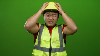 Sarı kasklı ve yelekli bir adam, parlak yeşil arka planda yüzünü buruşturuyor, elleri başının üstünde stres yapıyor..