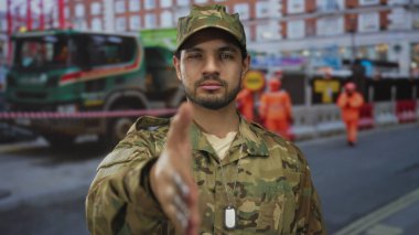 Kamuflaj üniformalı bir adam işaret parmağıyla inşaat alanındaki bariyerin yanındaki kamerayı işaret ediyor..