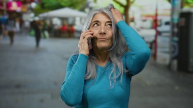 Gri saçlı yaşlı bir kadın kalabalık bir caddenin dışında duruyor, bir telefon görüşmesi yapıyor, günlük yaşamını ve iletişimini gösteriyor..
