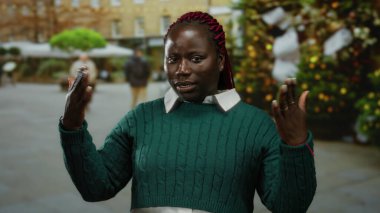 Güneşli bir şehir sokağında, yeşil bir süveter giyen, şenlikli bir arka planı olan bir kadın, kış mevsiminde sıcak ve kentsel bir hayat sürüyor..