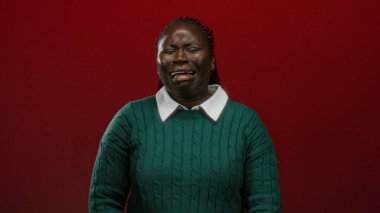 Yeşil kazaklı Afrikalı Amerikalı kadın, izole edilmiş kırmızı bir arka plana karşı duygusal bir şekilde ağlıyor. Üzüntü ve kırılganlık taşıyor..