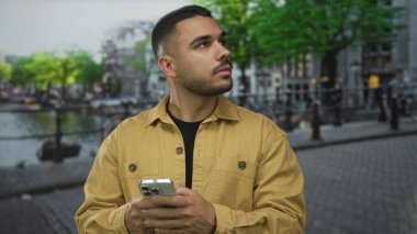 Sakallı, sarı ceketli adam akıllı telefonu tutuyor ve kanalın yanındaki sokağa bakıyor..
