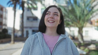 Açık havada gülümseyen esmer saçlı, gri kapüşonlu ve pembe gömlekli bir kadın güneşli bir sokakta, arka planda palmiye ağaçları olan..