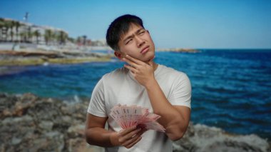 Tayvan banknotlarını ihtiyatlı bir şekilde deniz kenarında tutan genç Çinli bir adam. Arka planda okyanus dalgaları. Mali konuları çağrıştırıyor ve sahilde tefekkür ediyor..