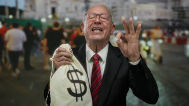 Takım elbiseli kıdemli adam elinde dolar sembolü olan para çantası tutuyor ve kalabalık bir sokakta iyi bir jest yapıyor..