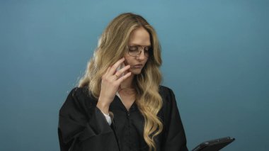 Sarışın bir kadın üniformalı bir yargıç, elinde telefon ve tablet, ciddi ve odaklanmış görünüyor, profesyonelliği ve otoriteyi simgeliyor..