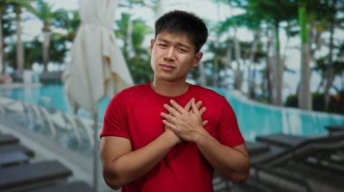 Kırmızı tişörtlü genç adam, havuzun yanında duruyor. Sakin bir açık hava tesisinde. Etrafı palmiye ağaçları ve boş şezlonglarla çevrili..