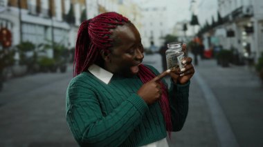 Dolarlarla dolu kavanozu elinde tutan kadın açık havada gülümsüyor para biriktirerek mutluluğunu gösteriyor Afro-Amerikan kadın şehir ortamında finans zevkini çıkarıyor.