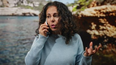 Sahil kenarında kıvırcık saçlı, bulanık arka planı ve berrak mavi suyu olan, sakin bir açık hava sahili tasvir eden bir kadın telefonda konuşuyor..