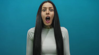 Şaşırdığını ifade eden bir kadın, ağzı açık ve elleri yüzüne yakın, izole edilmiş, canlı, mavi bir arka plana karşı..