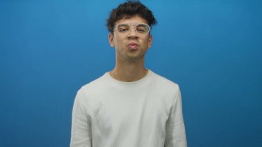 Koyu kıvırcık saçlı ve gözlüklü İspanyol adam mavi stüdyoyu işaret ediyor; eğlence enerjisi yaratıcılığı.