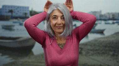 Kır saçlı yaşlı kadın sahilde teknelerin yanında şakacı kulak hareketleri yapıyor, uzak binaları olan sakin bir deniz kıyısına karşı neşeli kişiliğini sergiliyor..