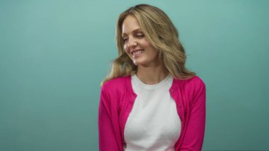 İzole edilmiş yeşil bir arka plan duvarının önünde gülümseyen bir kadın beyaz gömlek ve pembe kazak giyen kendine güveni ve sıcacık sarı saçları omuzlarının üzerinden dökülen bir kadın..