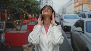 Şehir caddesinde beyaz laboratuvar önlüğü giyen kadın, şehir arabaları ve ağaçların arasında yoğun çevrenin altını çizerek hayal kırıklığı ve yorgunluk gösteriyor..