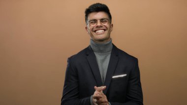 Genç bir adam kendine güvenerek gülerek ve alkışlayarak izole edilmiş kahverengi bir arka plana karşı şık giyiminin ve pozitif davranışının altını çiziyor..