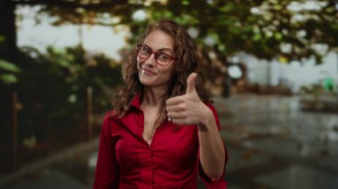 Kırmızı gözlüklü genç bir kadın parkın arka planında kırmızı tişört giymiş ve güneşli bir günde güvenle gülümsüyor.