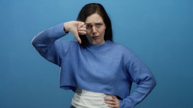 Mavi kazak giyen kadın, yüz ifadesi ve el hareketi yoluyla memnuniyetsizlik ya da hoşnutsuzluğu ileterek, sağlam bir mavi arkaplana karşı baş parmak işareti yapıyor..