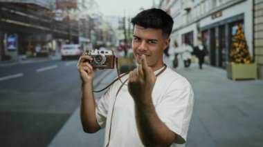 Elinde klasik kamera mimikleri olan genç bir adam açık şehir ortamında gündüz vakti işlek şehir caddesinde davetkar bir şekilde el kol hareketi yapıyor.