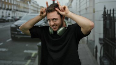 Sakallı ve kulaklıklı genç adam şehir caddesinde boynuz hareketi yapıyor. Arka planda arabalar ve cadde boyunca uzanan binalar var..