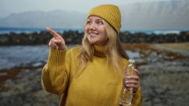 Sarı kazaklı ve şapkalı kadın deniz kenarına neşeli bir ifadeyle bakarken şişeyi tutuyor, plaj manzarasına karşı duruyor, rahat bir açık hava anı yakalıyor..