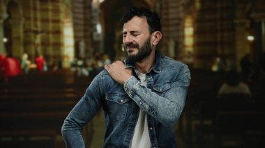 Kot ceketli, sakallı bir İspanyol Katolik kilisesinde duruyor ve omzunu tutarken rahatsız olduğunu söylüyor..