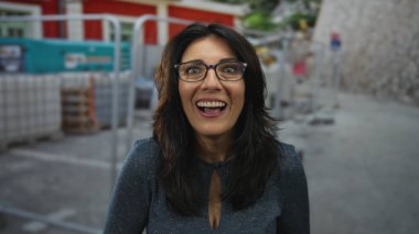 İnşaat alanında, etrafı inşaat iskelesi ve makinelerle çevrili, gözlüklü ve desenli mavi bluzlu gülümseyen bir kadın. Beklenmedik bir sürpriz..