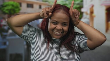 Çizgili gömlekli kadın kameraya bakıyor ve işaret parmağıyla caddeye işaret ederken gülümsüyor; neşe dolu.