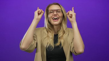 Kadın gülümsüyor ve iki eliyle yukarı doğru işaret ediyor canlı mor bir arka plan, gözlük ve bej bir ceket giyiyor, enerjik ve neşeli bir an yakalıyor.