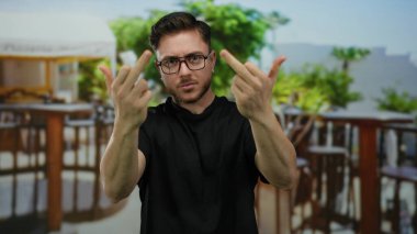 Sakallı genç adam şehir sokağının yanındaki terasta kaba bir jest yapıyor. Arka planda restoran ve kahve var..