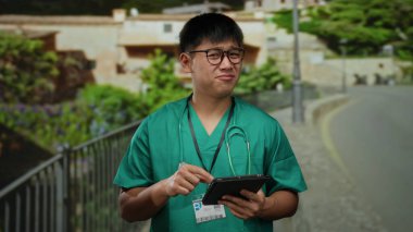 Tıbbi önlüklü genç adam şehir caddesinde tablet kullanıyor, boynunda steteskop ve yüzüne yansıyan güneş ışığıyla dikkatle odaklanıyor..