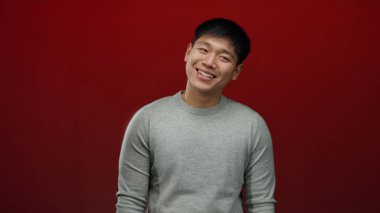 Gri süveter giymiş genç Çinli bir adam izole edilmiş kırmızı bir arka plan karşısında gülümsüyor. Mutluluk ve basitliği ifade ediyor..