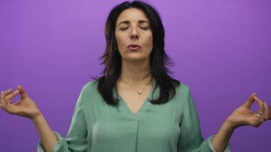 Yeşil bluzlu kadın stüdyoda mor duvara karşı kapalı gözleri olan Mudra 'yı tutuyor; farkındalık dinginliği.