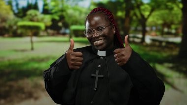 Yazar kıyafetli kadın, güneşli bir park ortamında başparmağını kaldırırken gülümsüyor, pozitifliğini ve sıcaklığını ifade ediyor..