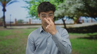 Gözlüklü genç adam yeşil parkta bulanık ağaçların arasında boyna dokunuyor ve bir yol; kişisel yansıma.