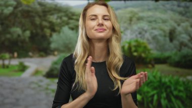 Güneşli bir günde, yemyeşil bir parkta, parlak bir gülümsemeyle, sarı saçlarını ve günlük kıyafetlerini sergileyen bir kadın..