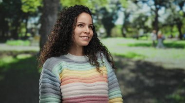 Açık havada güneşli bir günde arka planda ağaçlarla parkın tadını çıkaran renkli kazaklı bir kadın..