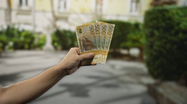 Şehrin arka planında bulanık bir sokakta Norveç kronu banknotları tutan bir adam, şehir ortamında açık hava mali işlemleri tasvir ediyor, döviz kurunu vurguluyor.