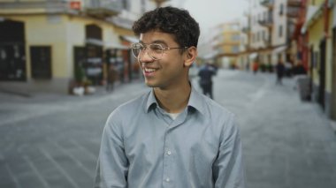 Gözlüklü genç İspanyol adam geniş bir gülümseme takınır ve hareketli bir şehir caddesinde dişlerini gösterir; mutluluk.