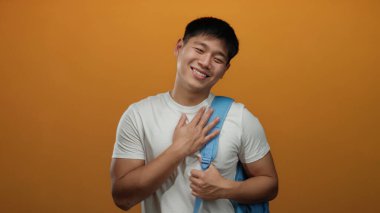 Genç adam, elleri turuncu duvar arkasında mutlu bir şekilde gülümsüyor. Beyaz gömlek ve mavi sırt çantasıyla duygusal jest ve pozitiflik sergiliyor.