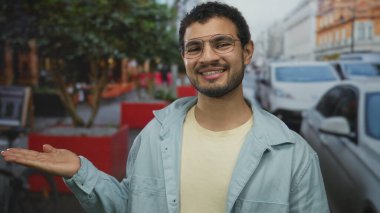 Şehrin hareketli caddesinde park etmiş arabaları ve kırmızı saksılarıyla açık el hareketleriyle İspanyol genç adam iyimserlik.