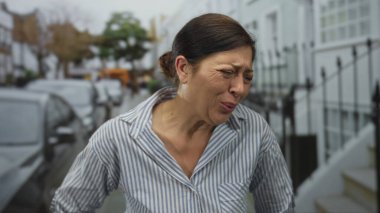 İspanyol orta yaşlı, çizgili tişört giyen kadın, park halindeki arabaların yakınındaki şehir sokağında belini tutuyor; acı.