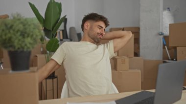 Adam yeni bir binada kollarını ve boynunu uzatıyor. Karton kutular taşınıyor ve tahta bir masada dizüstü bilgisayar var..