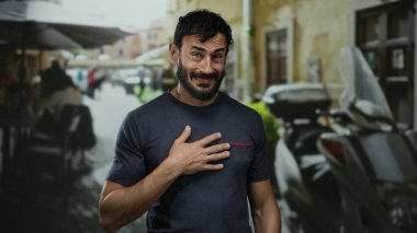 Sokaktaki İspanyol bir adam, motosiklet ve insanlarla dolu şehir sahnesinin arka planına karşı teşekkürlerini ve sakallarını ifade ederek minnettarlığını gösteriyor..