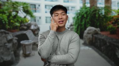 Genç adam, yeşil bitkiler ve modern binalarla dolu bir caddede yürürken akıllı bir telefonla konuşuyor, dinamik bir şehir manzarasında şehir yaşamını gözler önüne seriyor..