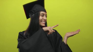 Mezuniyet elbisesi giymiş bir kadın, izole edilmiş sarı bir arka plan üzerinde avucunu gösteriyor, neşe ve akademik başarının somutlaşmış hali..