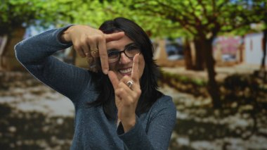Gülümseyen İspanyol kadın, yeşil bir park yolunda, yapraklı ağaçların altında elleri ve gözlükleriyle sahneyi çerçeveliyor; yaratıcı görüş.