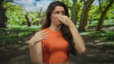 Yeşil parkta turuncu kolsuz bluz giyen genç İspanyol kadın güçlü bir açık hava kokusunu engellemek için burnunu çimdikliyor..