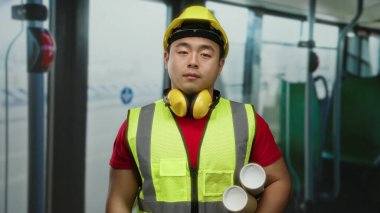 Güvenlik yeleği ve kasketi takmış, şehir gelişimini ve toplu taşıma planlarını sembolize eden Asyalı genç bir adam otobüsün içinde duruyor..