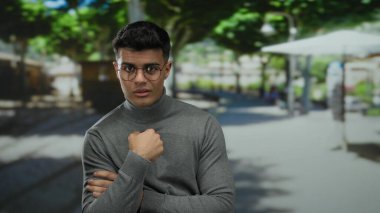 Gözlüklü genç adam güneşli bir şehir sokağında, açık şehir ortamında yeşil ağaçlarla çevrili neşeli bir ifadeyle kendini işaret ediyor..