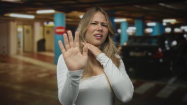 Otoparktaki sarışın kadın park halindeki bir arabanın yanında eli ile ağzını kapatıyor gözleri korkuyla genişlerken; şok paniği.